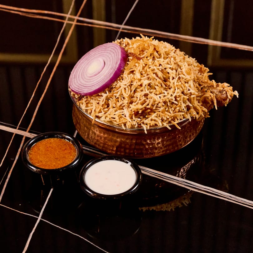 Chicken biryani in a copper bowl, garnished with red onion, served with two sauces.