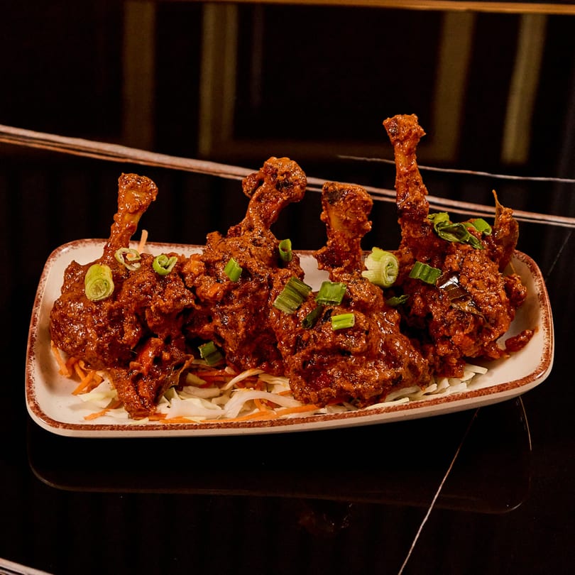 Four Indian chicken lollipops, coated in a rich red sauce, garnished with green onions on a bed of shredded cabbage.