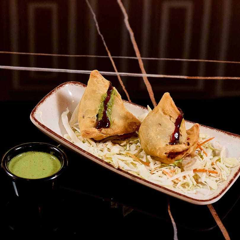 Two golden-brown samosas drizzled with green and brown sauces, served on shredded cabbage with a side of green chutney.
